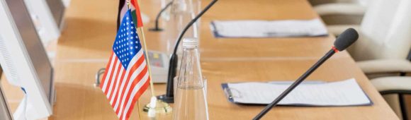 Row of bottle with water, national flags, microphones, computer monitors and papers on long table in conference hall
